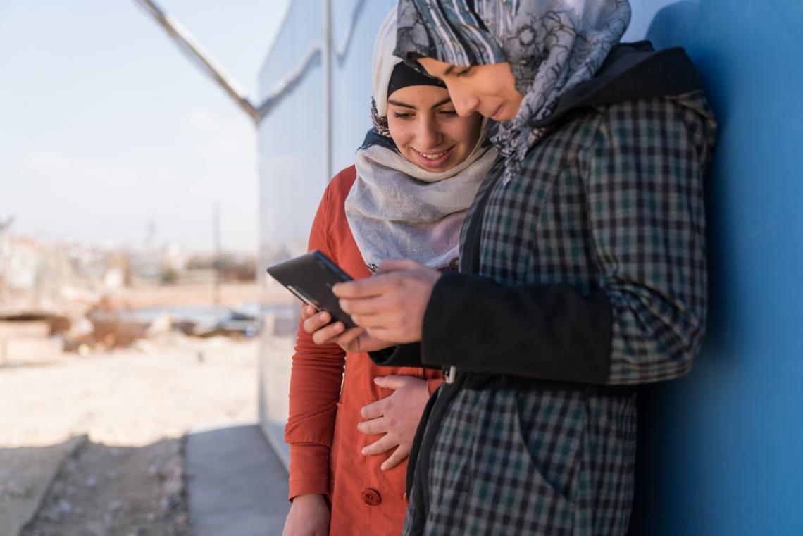 Two adolescent girls use a cellphone outside a solar kiosk in the Za’atari camp for Syrian refugees,